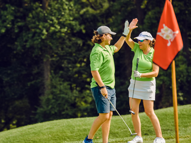 A man and woman high five on the green of a golf course. They are standing behind the hole, which is marked by a red flag with a shield that shows the pattern of Maryland's flag.