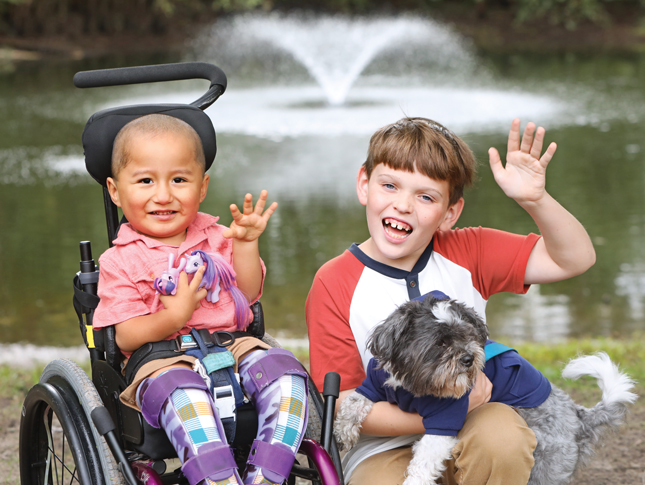 Posed photo of a young boy sitting in a wheelchair and a slightly older boy holding a small dog. Both boys are smiling and waving..