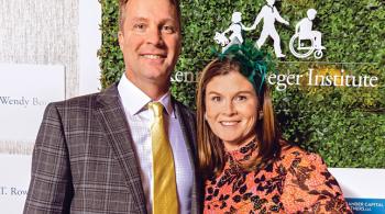 A husband and wife wearing fun formal wear pose for the camera, smiling. Behind them is a clear sign with Kennedy Krieger Institute’s logo on it. The sign hangs in front of a leaf-covered wall.