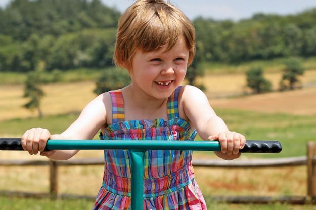 A young girl holds the handlebars of a teal tricycle. Behind her is a fence, with a hilly farm field on the other side.
