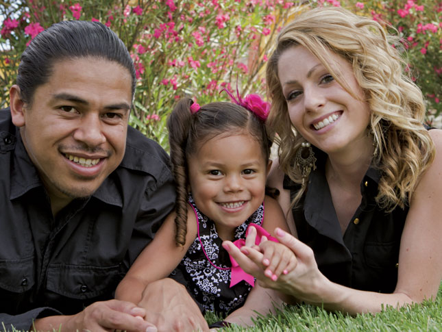 A father, daughter and mother lay in the grass and smile while facing the camera.