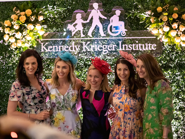 Women pose alongside a Kennedy Krieger banner at the Hats & Horses event