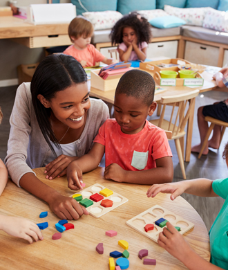 A teacher smiles as she helps three pre-school students with an activity at their desk.