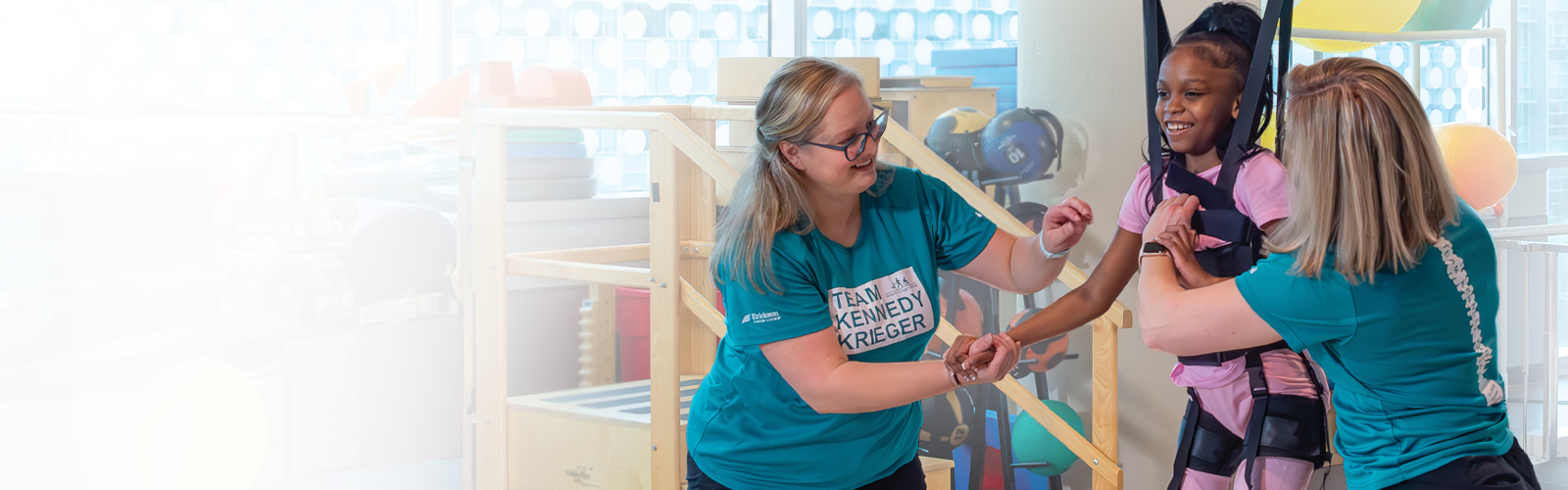 A young girl participates in physical therapy using assistive technology. Two female therapists are overseeing the session.
