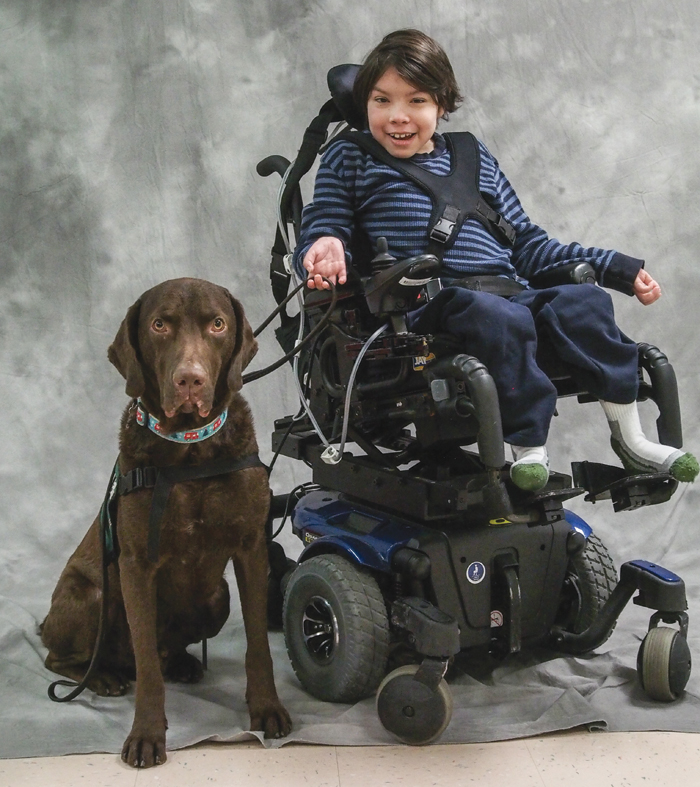 A child in an electric wheel chair smiling and sitting with a chocolate labrador.