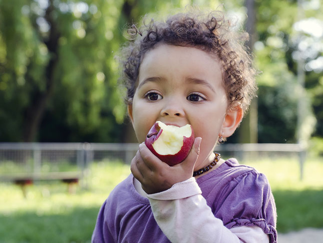 A young boy eats a red apple while standing outside.