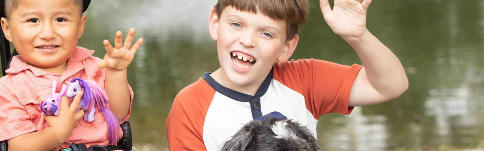 Two boys sit next to each each other and smile while waving at the camera. The boy on the left is sitting in a mobility device and is holding two toys. The boy on the right is sitting on the ground is holding a small dog.