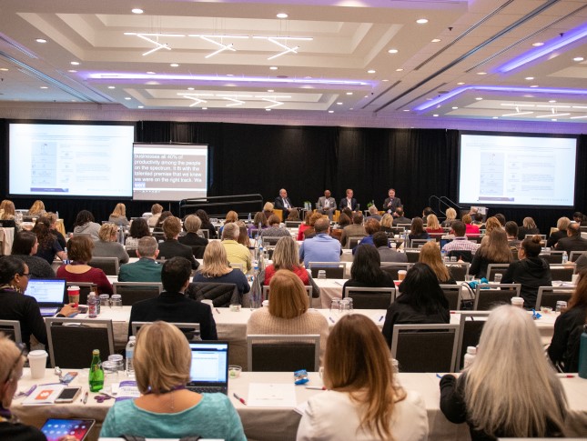 A seated crowd watches a panel of speakers in a large conference room.