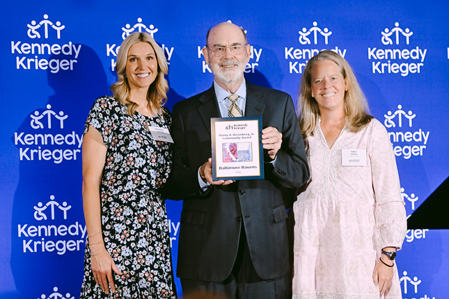 Three people stand in front of a Kennedy Krieger step-and-repeat backdrop. The person in the center holds a framed award plaque that reads Henry A. Rosenberg, Jr. Community Award with Baltimore Ravens visible at the bottom, while the two people on either side pose beside the recipient.