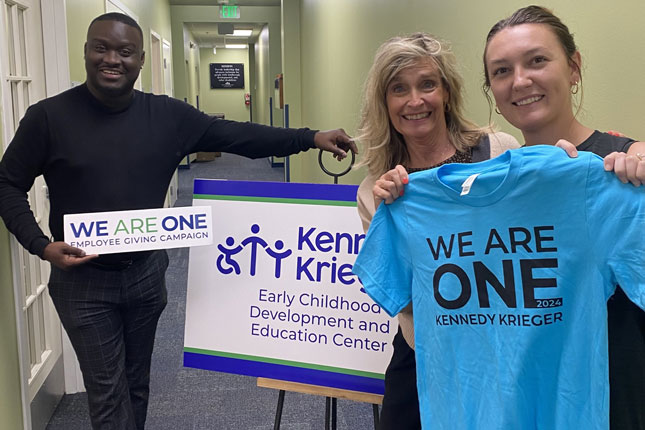 A man and two women smile while standing on either side of a Kennedy Krieger Early Childhood Development and Education Center sign that is displayed on an easel. The man is standing on the left holding a We Are One sign, while one of the women on the right is holding a We Are One 2024 t-shirt.