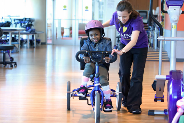 A physical therapist assists a young girl riding a tricycle. 