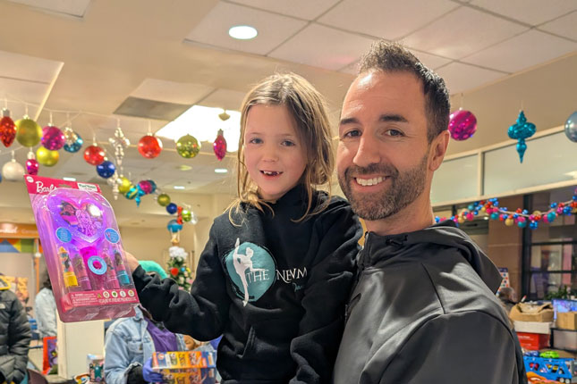 A father holds his daughter, who holds a Barbie toy set. Behind them are holiday decorations and piles of other toys. They are indoors in what could be a conference room or lobby.