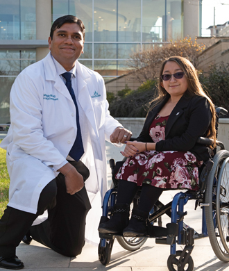 A doctor kneels next to a patient in a wheelchair at Kennedy Krieger's Therapy Garden. Both are looking at the camera and smiling.