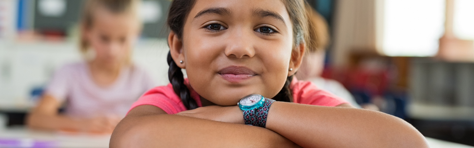 A young girl sits at her school desk with a slight smile. 