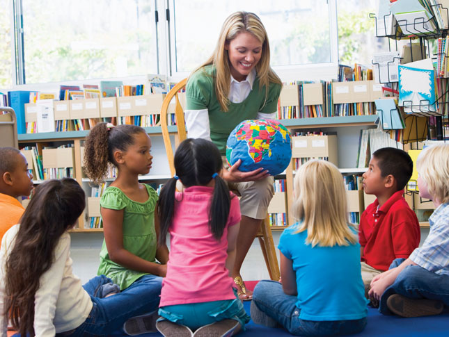 A teacher shows her students a globe in a classroom. She is sitting on a chair smiling, with her students sitting in front of the chair studying the globe.
