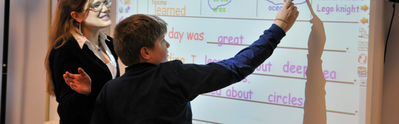 A boy writes on a whiteboard while his teacher, a woman, stands to his left.