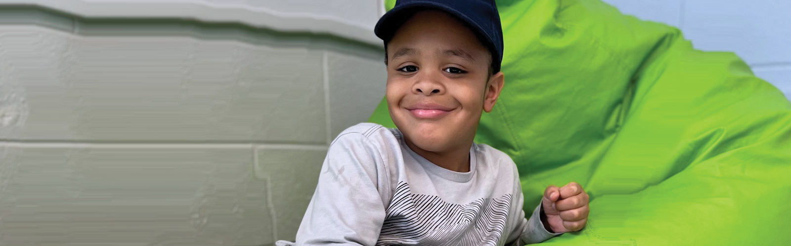 A young boy sits against a bright green beanbag chair and smiles.