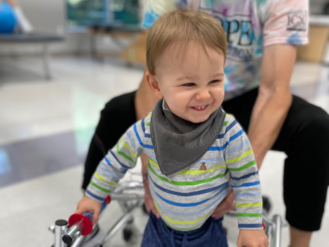 A little boy smiles while using a walker during a physical therapy session.