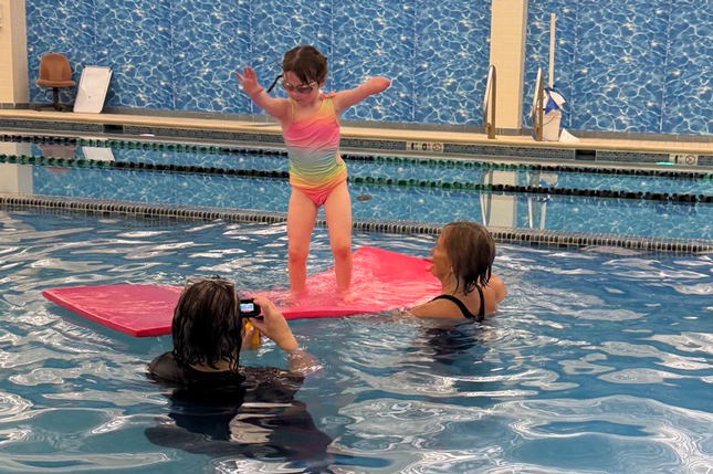 Marion Durfee stands on a paddle board in a pool as a coach and another adult look on.