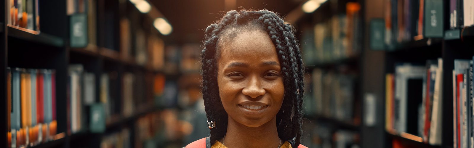 A young black woman stands in the aisle of a library, with books visible on the bookshelves behind her.