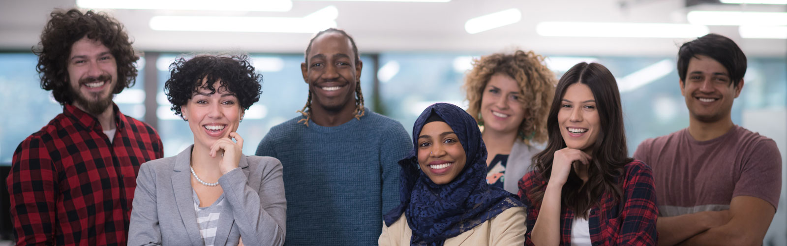 A diverse group of seven young professionals, three men and four women, smile at the camera.