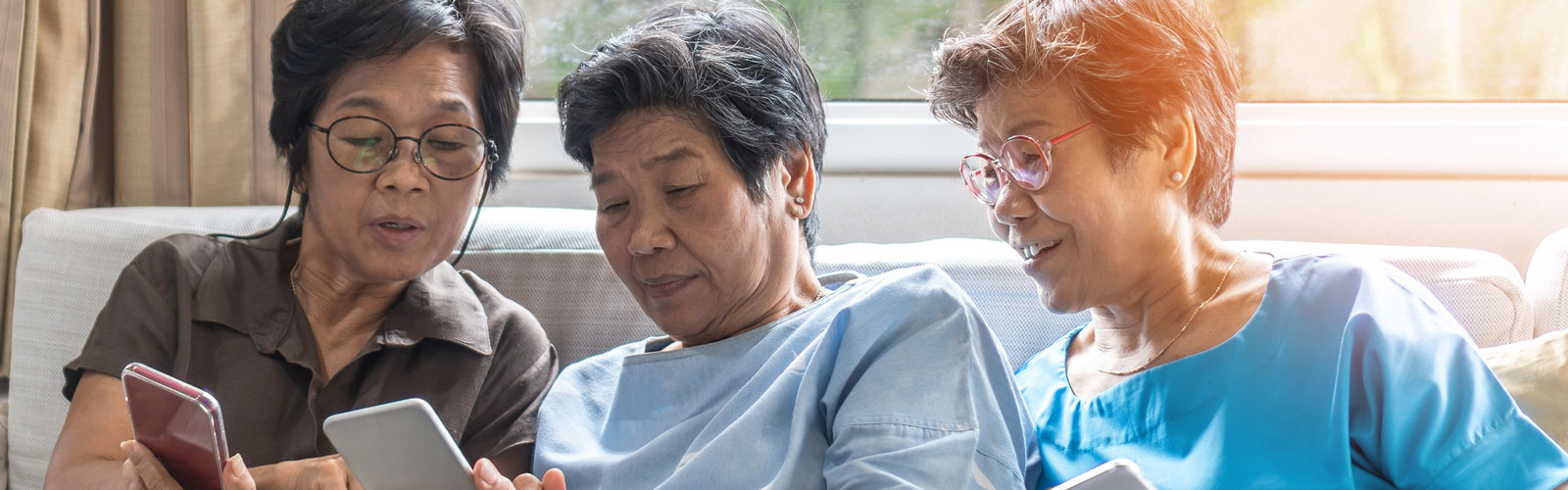 Three elderly women sitting next to each other on a sofa, using smartphones.