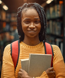 Young Black woman stands in the aisle of a library holding books. 