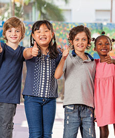 Four children standing shoulder to shoulder smiling, with thumbs up.