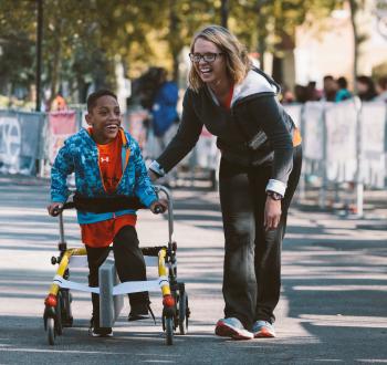 A child using a walker with assistance from an adult in a sport program, both smiling brightly.