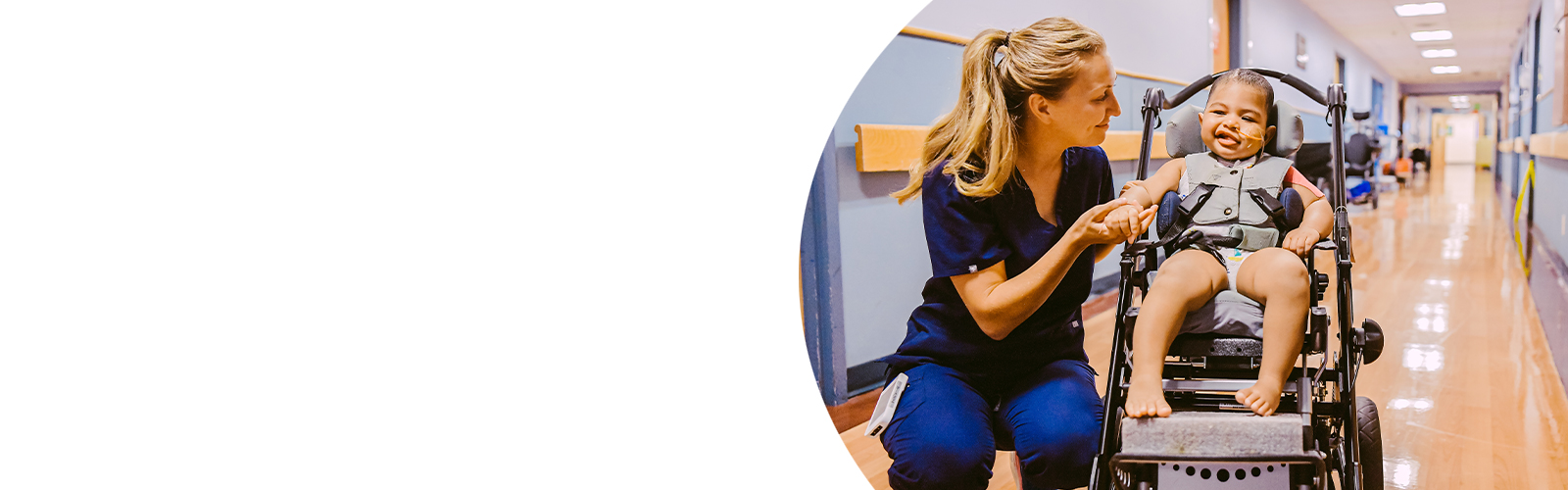 A nurse kneels to the side of a young boy in a wheelchair and looks at him. They are both smiling.