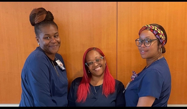 Three smiling nurses stand against a wall for a group photo.