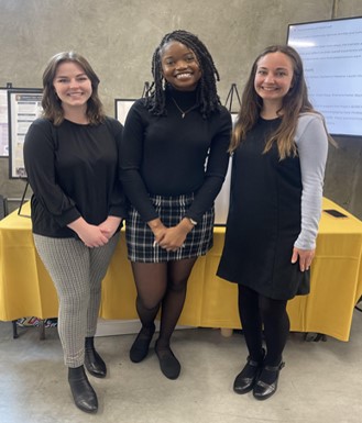 Three young women stand in front of a table with a yellow tablecloth.