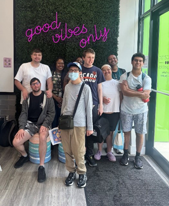A group stands in front of a wall with an artificial green surface, with a sign that reads Good Vibes Only mounted on the surface. The sign is pink and in script font