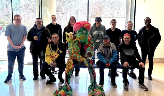 A group photo in the lobby of the Baltimore Museum of Art. The photo is staged around a bench with a colorful sculpture, with some sitting next to the sculpture and others standing behind it