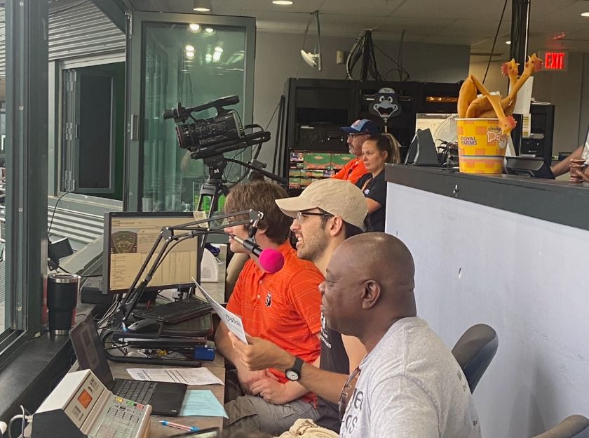 Robin talks into a microphone inside of a press box at baseball game. He is sitting between two men.