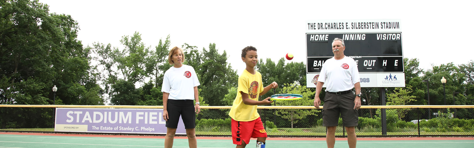 A boy bounces a tennis ball off his racket, while his coaches--a woman and a man--stand behind him and watch.