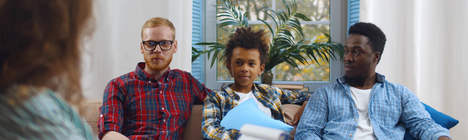 A therapist talks to a couple and their son. The therapist, a woman, has her back to the camera while two fathers and a son sit on the sofa facing the camera.