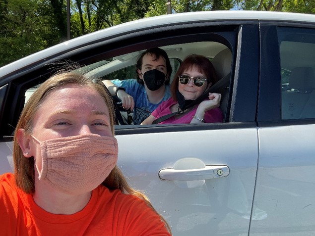 A woman wearing a face mask and an orange t-shirt takes a selfie with a man and a woman sitting in a car behind her.
