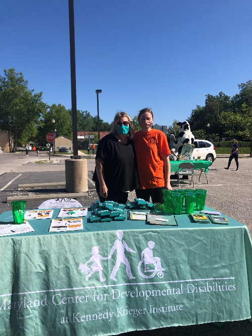 Two women wearing face masks, one wearing a black shirt and another wearing orange, stand behind a Kennedy Krieger Institute branded table. 