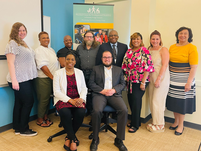 A group photo from a Faith Community meeting. A woman and man sit in the front with a group of four people standing behind them. The top of a green and yellow MCDD banner can be seen in the very back.