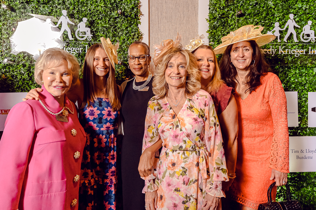 Six women stand for a photo at Hats & Horses 2023. Behind them are two green backdrops with the Kennedy Krieger logo in the middle.