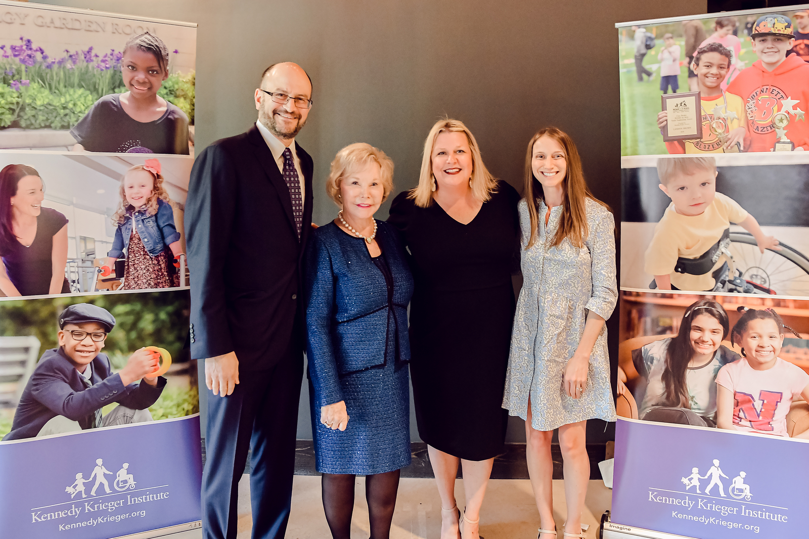 Standing left to right: Dr. Bradley Schlaggar, Kennedy Krieger Board chair Nancy Grasmcik, MCDD director Maureen van Stone and Mallory Legg. They are between two Kennedy Krieger banners at the Society Party.