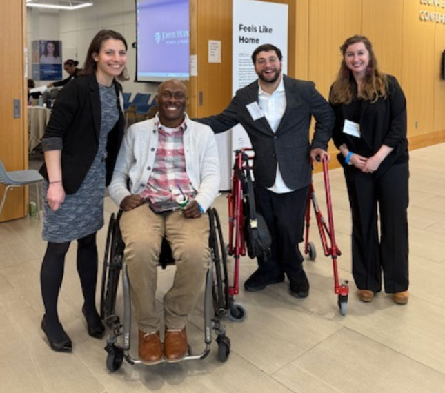 Chris Mason-Hale in a group photo with two women and a man at the Opening Doors to Home Accessibility workshop. The two women are standing on the ends, while Mason-Hale sits second to the left in a wheelchair. The man next to him is using a walker.