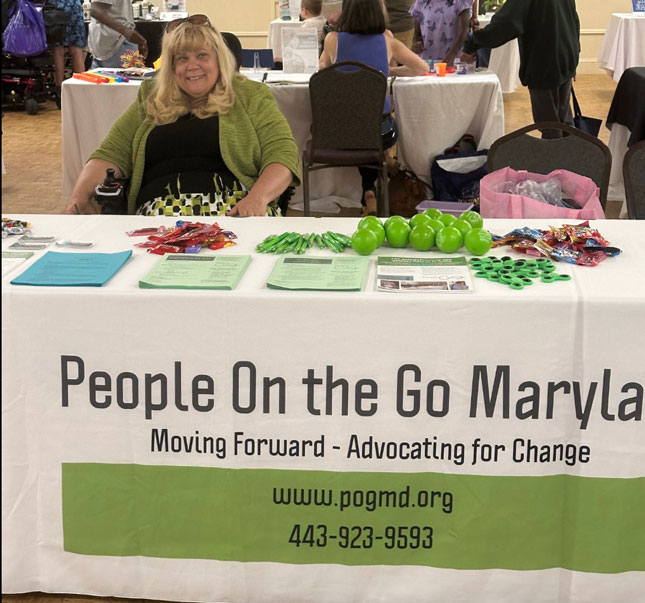 Tracy Wright sits at People on the Go Maryland's exhibitor table. The is covered by a cloth with People On the Go Maryland's branding, with flyers and other items sitting on the cloth.