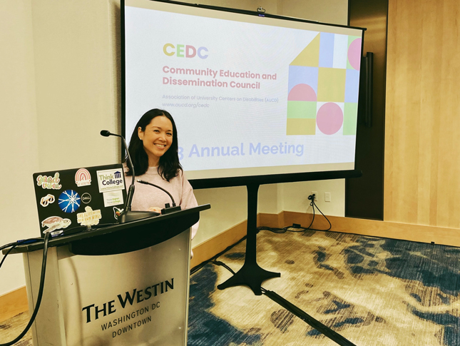 Kristine Nellenbach stands a podium and smiles during the annual meeting for AUCD’s Community Education and Dissemination Council (CEDC). 