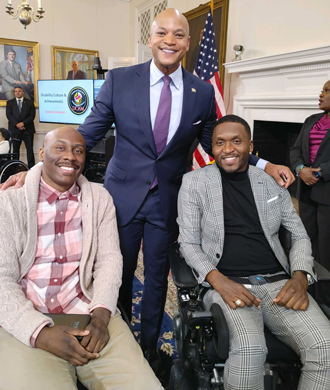 Chris Mason-Hale, Maryland Governor Wes Moore and another man smile in a photo. Mason-Hale and the other man are sitting in their wheelchairs, while Governor Moore stands behind them and smiles. 
