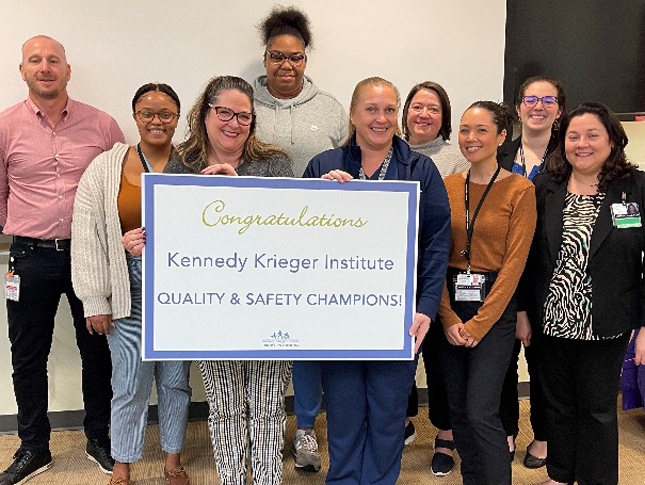 A group of people smile for a photo, with two of them in the front each holding one side of a large sign that reads "Kennedy Krieger Institute Quality & Safety Champions!"