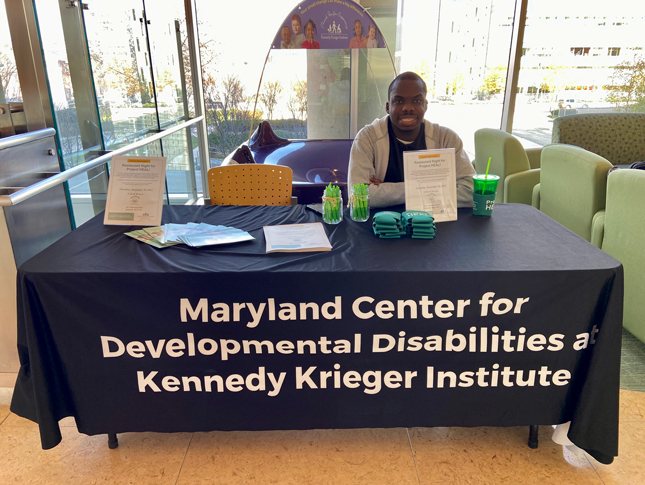 Ugo smiles while sitting at a table. The table is covered with a blue table cloth that says Maryland Center for Developmental Disabilities at Kennedy Krieger Institute in the front, with promotional options on the top.