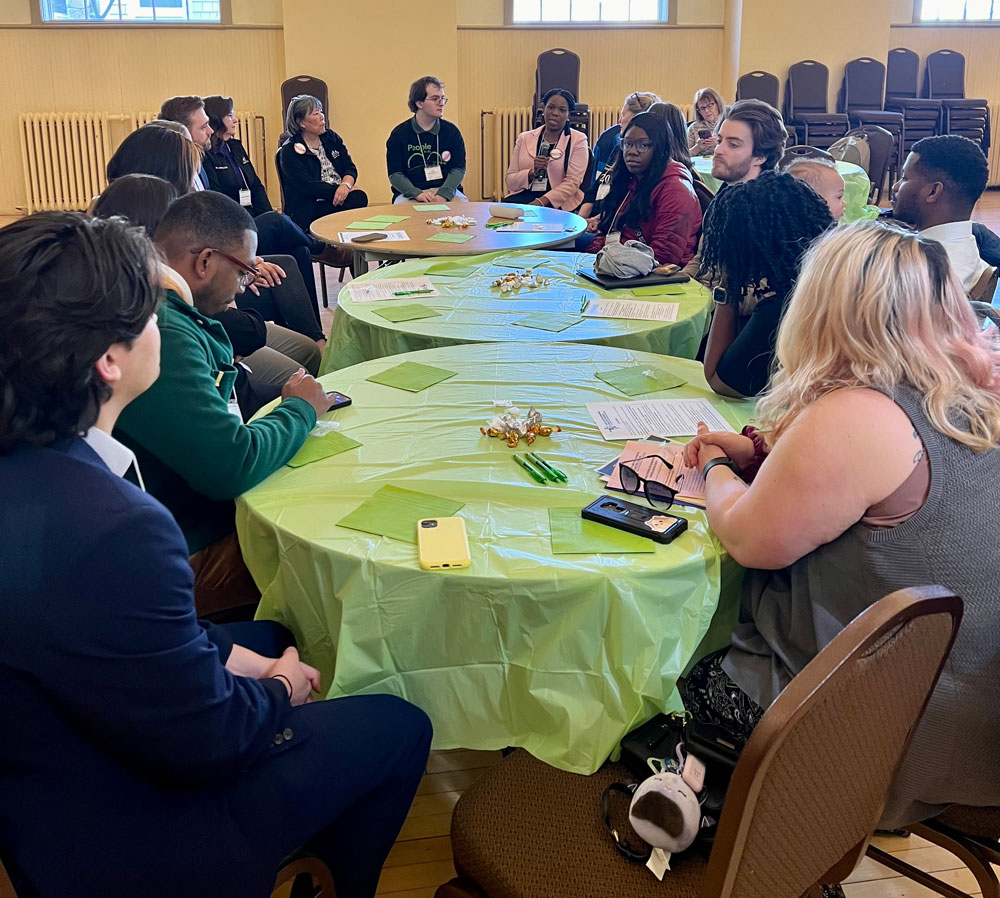 A large group of people sit around three round tables that have been pushed together. 