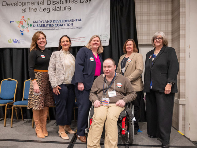 Five women stand behind Mat Rice and smile for a group photo. Rice is seated in his wheelchair. The group is in front of a Developmental Disabilities Day at the Legislature banner.